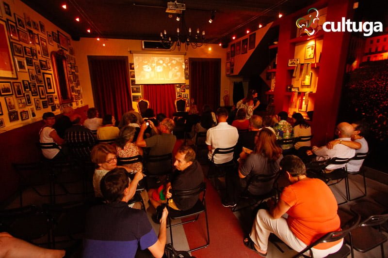 Público aguarda a entrada dos músicos do Fado ao centro em uma sala com muitas fotos e luz baixa